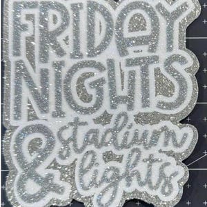 May include: A silver glitter and white sticker featuring the text "FRIDAY NIGHTS & stadium LIGHTS". The words are in a layered design with silver glitter outlining the text. The sticker has an irregular, unique shape, with a sparkling effect.