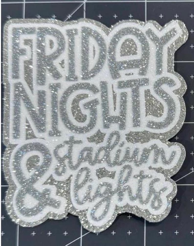 May include: A silver glitter and white sticker featuring the text "FRIDAY NIGHTS & stadium LIGHTS". The words are in a layered design with silver glitter outlining the text. The sticker has an irregular, unique shape, with a sparkling effect.