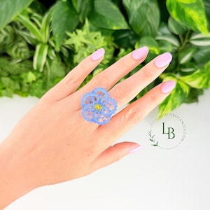Könnte beinhalten: Ein Ring mit einem hellblauen, filigranen, spitzenartigen Blumenmuster und einem kleinen, leuchtend gelben Stein in der Mitte. Das zarte, durchbrochene Design bildet eine kreisförmige, blütenblattartige Form, die auffällig auf dem Finger sitzt.