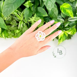 May include: A white lace ring featuring a delicate floral pattern with a small, bright yellow center accent. The intricate, circular lace design is centered on the band, creating a detailed, textured appearance against the background.