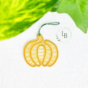 Peut inclure: Une décoration en dentelle jaune en forme de citrouille avec un travail de fil complexe et une boucle verte sur le dessus pour la suspendre. Le motif inclut un logo circulaire avec le texte LB Lacybotanics imprimé à droite sur un tissu blanc.