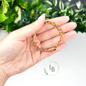 May include: A multi-coloured braided bracelet in red, yellow, green, and purple. The bracelet is held in a hand, with a white background and green foliage in the background. The bracelet is knotted to secure it.