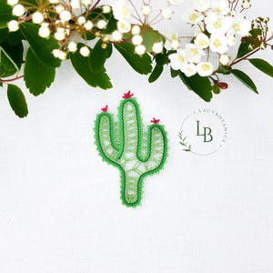 May include: A green cactus-shaped lace applique featuring intricate threadwork and three small pink floral accents at the top. The design includes a circular logo with the text LB LACYBOTANICS positioned to the right of the cactus.