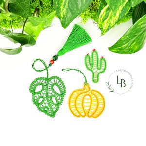 Puede incluir: Tres adornos decorativos de encaje que incluyen una hoja de monstera verde, una calabaza amarilla y un pequeño cactus verde con detalles rojos. Una borla verde con cuentas rojas y verdes está unida a la hoja. El texto LACYBOTANICS.COM es visible.