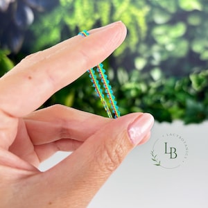 Può includere: Primo piano di un braccialetto colorato e delicato tenuto tra le dita. Il braccialetto presenta un motivo ripetuto di piccoli accenti turchesi e rossi. Lo sfondo è sfocato, con fogliame verde e il logo Lacy Botanics.