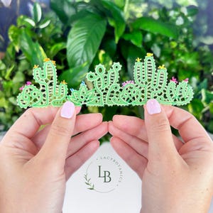 Könnte beinhalten: Eine hellgrüne Spitzenapplikation mit einem Wüstenlandschaftsdesign und mehreren Kakteenformen. Die aufwendige Stickerei enthält kleine, bunte florale Akzente aus gelbem, rosa und blauem Garn, die über die Kakteensilhouetten verteilt sind.