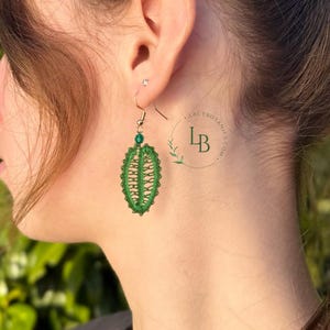 Puede incluir: Un pendiente verde con forma de hoja hecho de hilo fino, con un intrincado patrón calado y una pequeña cuenta verde en la parte superior. El pendiente cuelga de un gancho de metal. Se ve un logotipo circular con el texto LACYBOTANICS.COM.