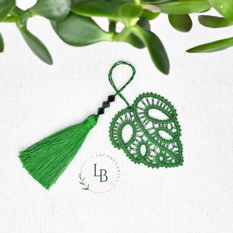 May include: A green lace monstera leaf ornament featuring a delicate thread-work pattern, a matching green tassel, and three dark faceted beads on a hanging loop. The logo LB Lacybotanics is printed in dark green text near the leaf ornament.