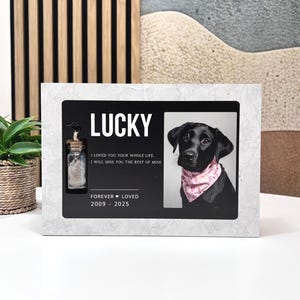 Può includere: Una cornice commemorativa con il nome "LUCKY" in lettere bianche. La cornice include una foto di un cane nero con una bandana rosa, una piccola bottiglia di vetro e il testo "I LOVED YOU YOUR WHOLE LIFE. I WILL MISS YOU THE REST OF MINE. FOREVER LOVED 2009 - 2025."