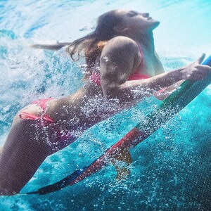 A vibrant underwater image showcasing a woman gracefully riding a teal surfboard. She's wearing a pink bikini and is partially submerged, creating a dynamic splash effect. The water is a clear turquoise, and the overall scene is energetic and action-packed, capturing the thrill of water sports.