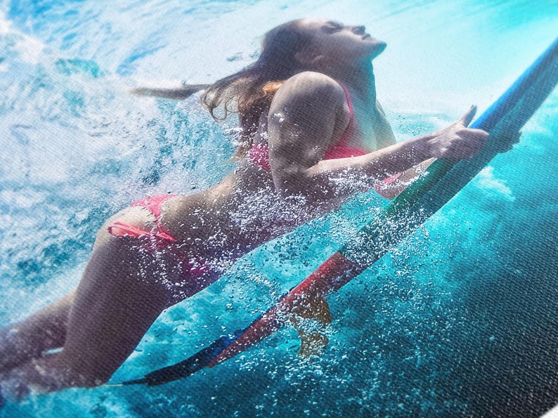 A vibrant underwater image showcasing a woman gracefully riding a teal surfboard. She's wearing a pink bikini and is partially submerged, creating a dynamic splash effect. The water is a clear turquoise, and the overall scene is energetic and action-packed, capturing the thrill of water sports.