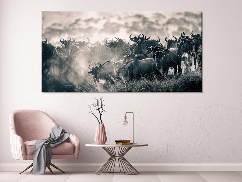 A panoramic monochrome photograph depicts a large herd of wildebeest in a sepia tone. The animals are densely packed, their forms partially obscured by dust and the low light. The wildebeest are predominantly dark, with some lighter areas suggesting highlights from the sun. The background is a hazy, light-colored sky, creating a contrast with the dark figures of the animals. The overall mood is one of movement and wildness, characteristic of wildlife photography.