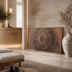 A rectangular wall art piece featuring a detailed brown and black wood grain pattern of tree rings, framed in light wood. The artwork displays concentric circular lines radiating from a central point, mimicking the cross-section of a tree.