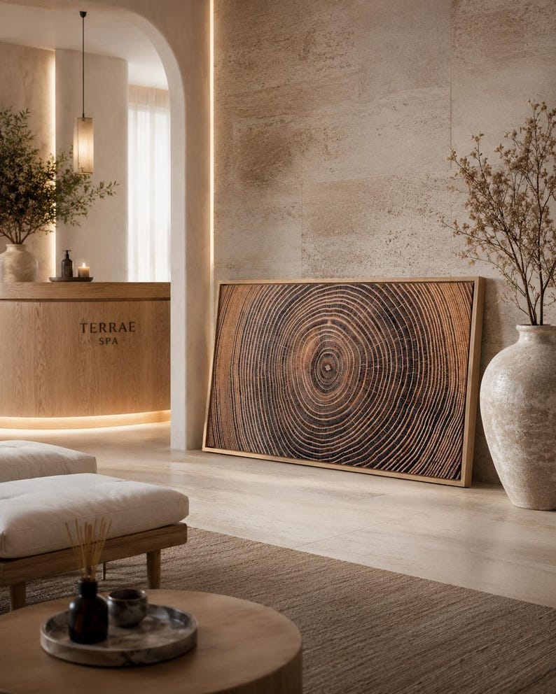 A rectangular wall art piece featuring a detailed brown and black wood grain pattern of tree rings, framed in light wood. The artwork displays concentric circular lines radiating from a central point, mimicking the cross-section of a tree.