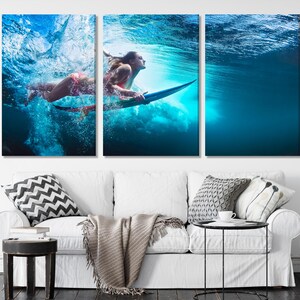 A three-panel triptych canvas print depicting a surfer underwater. The vibrant blue water is filled with bubbles and light. The surfer is positioned dynamically, creating a sense of motion and energy. The artwork features a woman in a bikini, surfing on a surfboard. The colors are predominantly blues and greens, with hints of pink from the surfer's attire. This artwork is perfect for adding a coastal vibe to any living space.