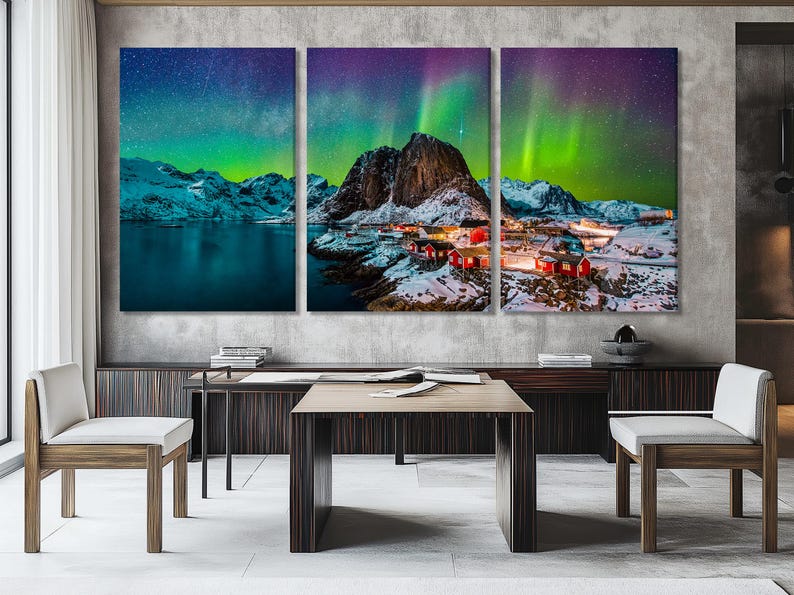 A snowy coastal mountain scene shows red cabin houses lit warmly below bright green aurora streaks across a dark sky over dark water
