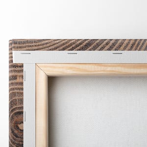 The back of a rectangular canvas print featuring a dark brown wood-grain frame. The white canvas is stretched over a light-colored wooden stretcher bar frame, secured with metal staples along the edge and a gold-toned sawtooth hanger.