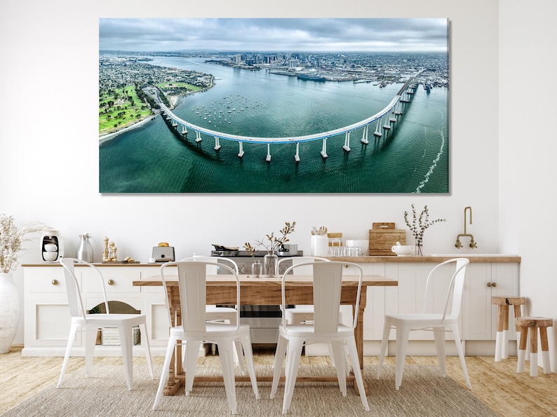 Aerial view of a curving bridge spanning teal water with a marina, city skyline, and cloudy sky