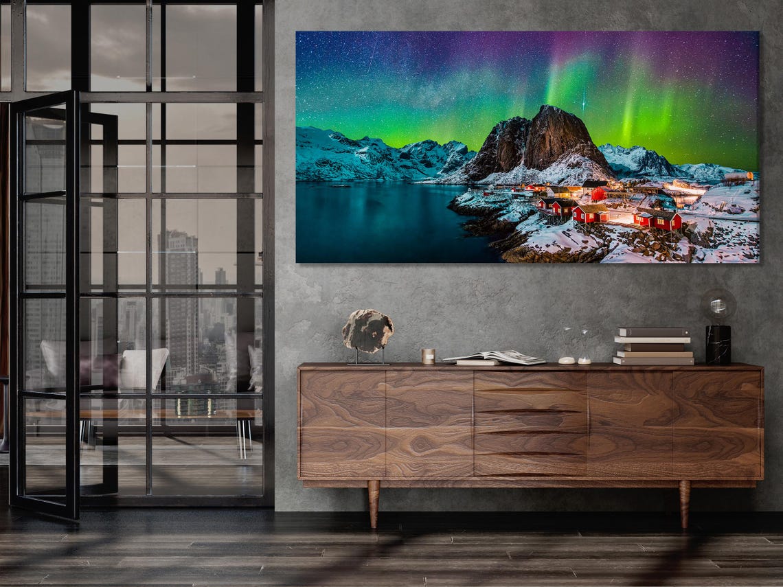 A snowy coastal mountain scene shows red cabin houses lit warmly below bright green aurora streaks across a dark sky over dark water detail view