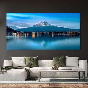 May include: A panoramic photograph of Mount Fuji, a snow-capped mountain, reflected in a still lake. The mountain is in the distance, with a town in the foreground. The sky is a vibrant blue with wispy clouds.