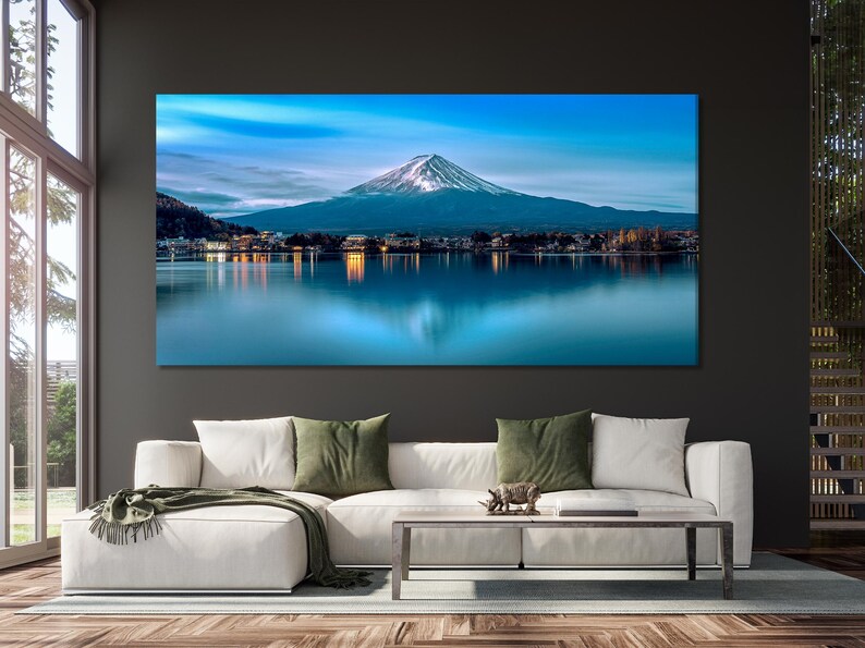 May include: A panoramic photograph of Mount Fuji, a snow-capped mountain, reflected in a still lake. The mountain is in the distance, with a town in the foreground. The sky is a vibrant blue with wispy clouds.