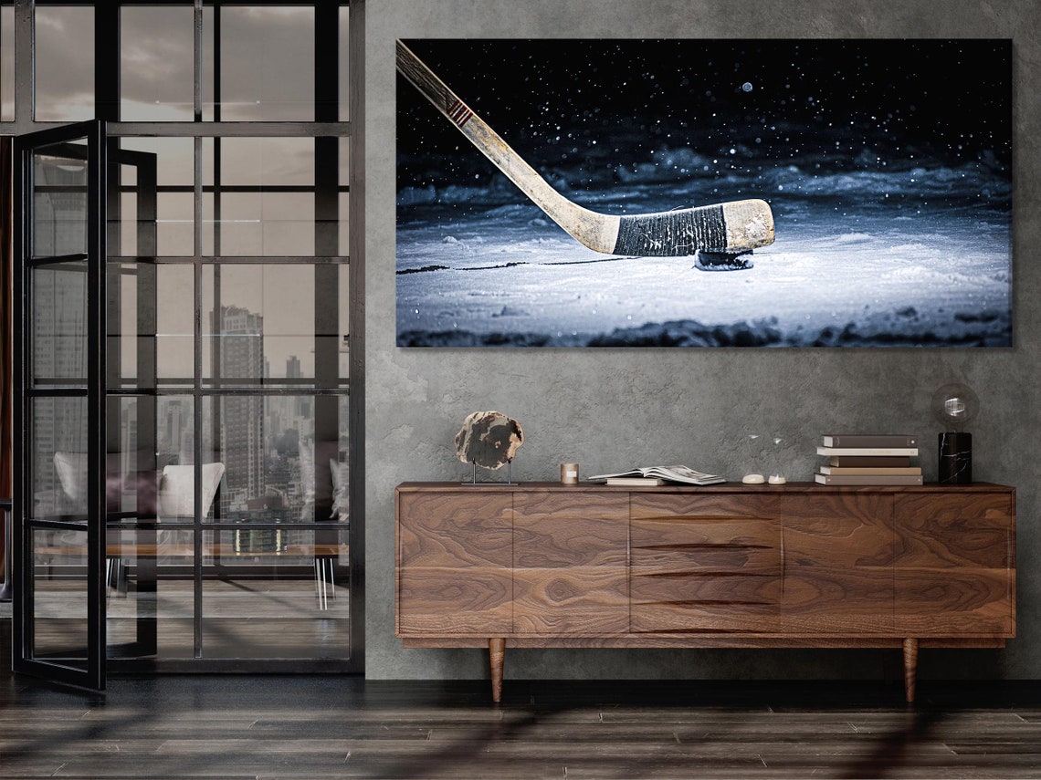 Snowy ice hockey scene with a hockey stick angled across the frame and a puck on bright ice, falling snow in a dark night sky detail view