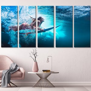 Five-panel canvas print depicting two women underwater, surfing on a surfboard. The vibrant blue water is filled with bubbles and sunlight. The women are wearing bikinis and are positioned dynamically on the board. The overall theme is aquatic sports and action photography. The image is suitable for home or office decor.