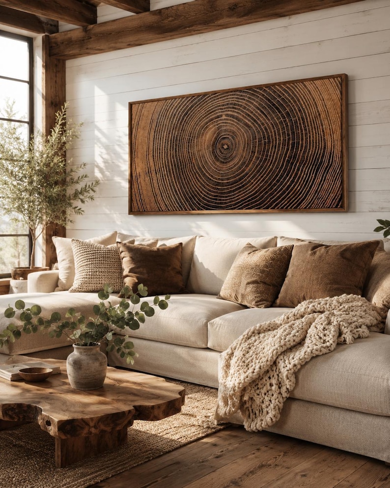 A rectangular wall art piece featuring a detailed dark brown wood grain cross-section with concentric rings, framed in wood. The artwork is displayed above a beige sectional sofa with brown and cream pillows and a chunky knit throw blanket.