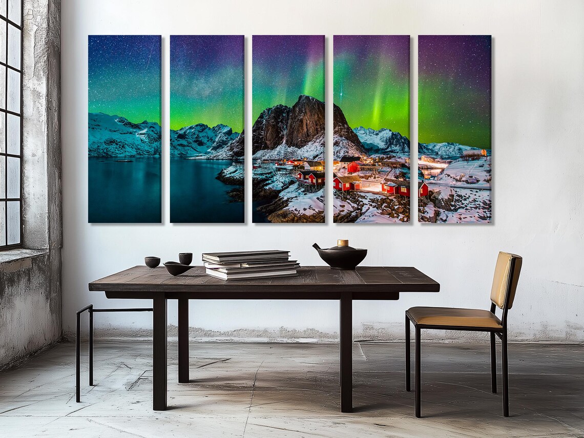 A snowy coastal mountain scene shows red cabin houses lit warmly below bright green aurora streaks across a dark sky over dark water detail view