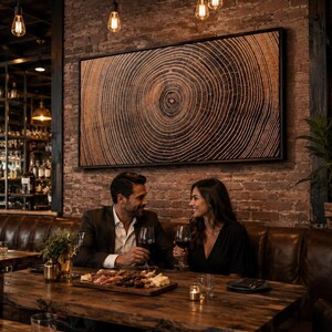 A large rectangular framed wall art piece featuring a detailed wood grain cross-section with concentric rings in shades of brown and black, mounted in a thin black frame on a rustic exposed brick wall above a leather booth.