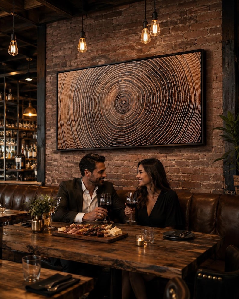 A large rectangular framed wall art piece featuring a detailed wood grain cross-section with concentric rings in shades of brown and black, mounted in a thin black frame on a rustic exposed brick wall above a leather booth.