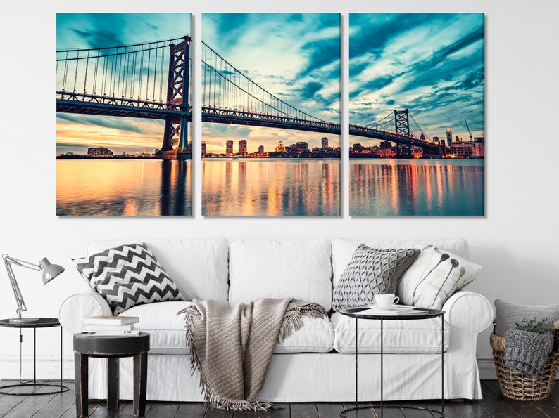 Three-panel suspension bridge cityscape at dusk with teal-blue sky and warm sunset reflections on water