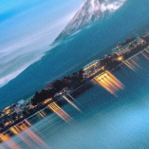 May include: A panoramic view of a mountain range with a lake in the foreground. The lake is illuminated by lights reflecting off the water. The mountain range is covered in snow and the sky is a light blue.