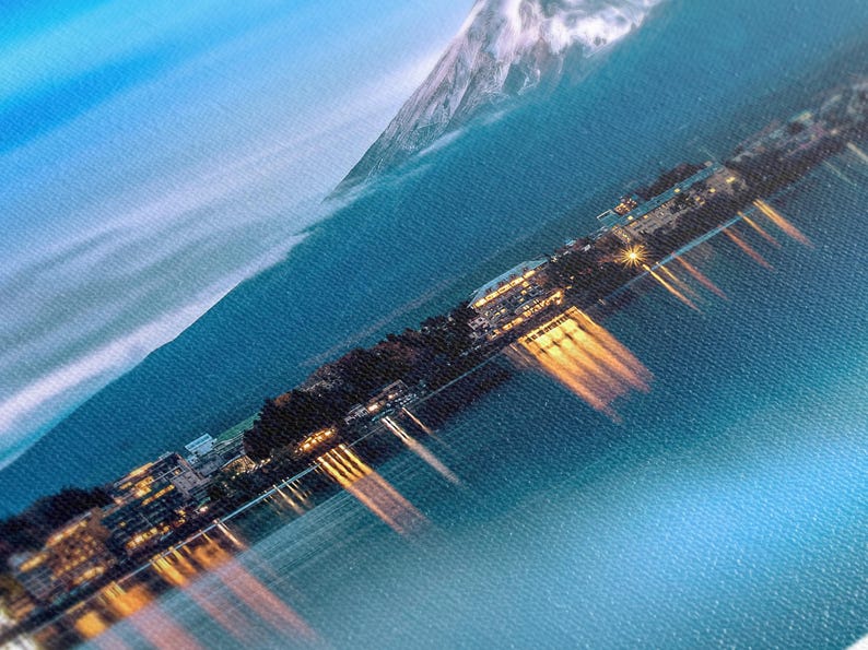 May include: A panoramic view of a mountain range with a lake in the foreground. The lake is illuminated by lights reflecting off the water. The mountain range is covered in snow and the sky is a light blue.
