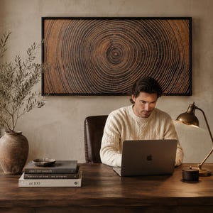 A rectangular wall art piece featuring a dark brown wood grain cross-section with visible concentric rings, framed in black. The artwork is displayed above a wooden desk with a laptop, a stack of three books, a small bowl, and a lamp.