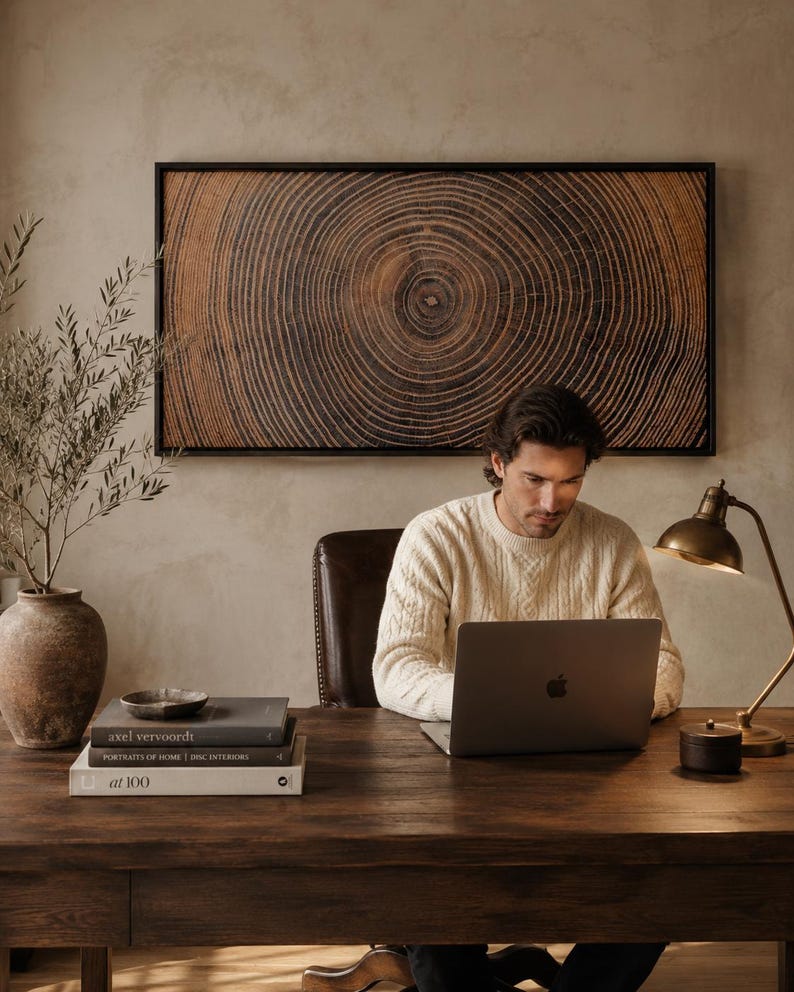 A rectangular wall art piece featuring a dark brown wood grain cross-section with visible concentric rings, framed in black. The artwork is displayed above a wooden desk with a laptop, a stack of three books, a small bowl, and a lamp.