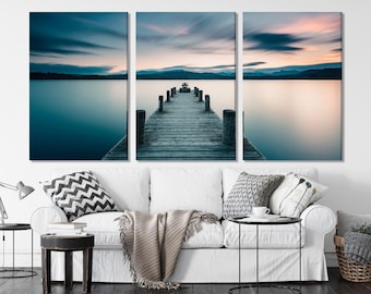 May include: A wooden jetty stretches out into a calm lake, with a cloudy sky and a mountain range in the distance. The jetty is made of weathered wooden planks and has a few wooden posts on either side. The water is a deep blue colour and reflects the sky.