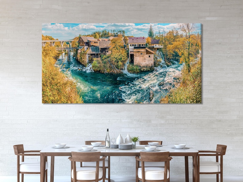 Wide river landscape with turquoise water, waterfalls, autumn trees and a village under a blue sky