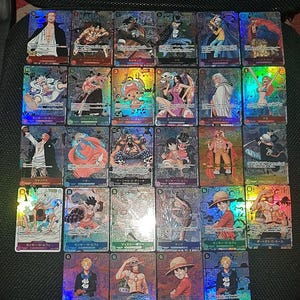 May include: A selection of holographic trading cards, each depicting anime characters with Japanese text. The cards are brightly coloured and arranged on a dark surface. The image highlights the cards' detailed artwork and collectable appeal.