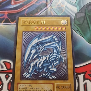 May include: A Yu-Gi-Oh! trading card depicting the Blue-Eyes White Dragon. The card features a blue dragon illustration on a gold background with Japanese text. Attack is 3000 and defence is 2500.