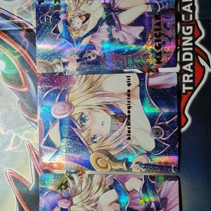 Può includere: Tre carte da gioco olografiche con un personaggio femminile con lunghi capelli biondi che indossa un abito viola e nero. Il personaggio tiene uno scettro con un globo dorato all'estremità. Le carte sono della serie "Black Magician Girl".