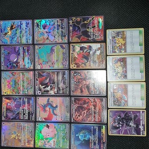 Può includere: Una collezione di carte collezionabili olografiche con vari personaggi e disegni. Le carte presentano illustrazioni colorate e testo, alcune con l'etichetta "Champions Festival". Le carte sono disposte su una superficie scura.