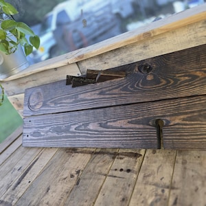 May include: A dark stained wooden wine glass and bottle holder. The holder is made of two planks of wood with a carved slot for the bottle and a circular indentation for the glass. The wood grain is visible.