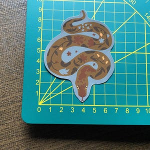 Może przedstawiać: Naklejka z brązowym i niebieskim wężem z kwiatowymi wzorami. Wąż jest zwinięty w kółko z ogonem w pysku.