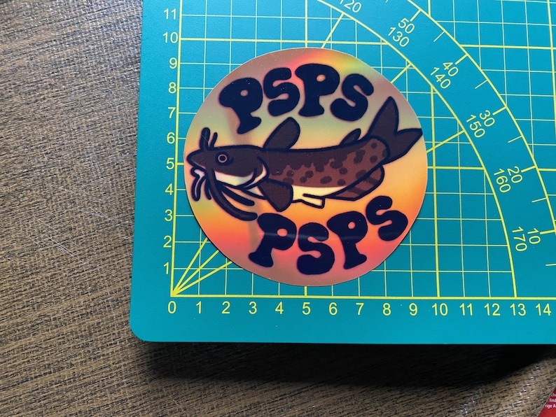 May include: A round sticker with a rainbow background and a black catfish with white spots. The sticker has the text "PSPS" in black letters on the top and bottom.