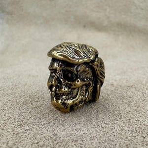 Pode incluir: Uma escultura detalhada de crânio em latão com uma superfície texturizada. O crânio está usando um chapéu com cabelo detalhado. A escultura é um item decorativo único.