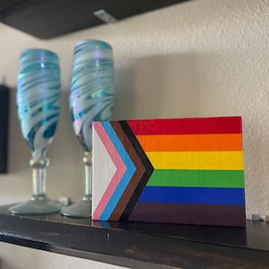 May include: A wooden sign with a painted version of the Progress Pride flag. The flag has the traditional rainbow colors with a black and brown stripe at the top, a light blue, pink, and white triangle at the left, and a dark brown stripe at the bottom.