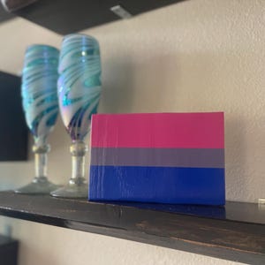 May include: A small rectangular artwork featuring the colors of the bisexual pride flag: pink, gray, and blue. Two decorative wine glasses with blue and white swirls are behind the artwork. The items are displayed on a dark shelf.