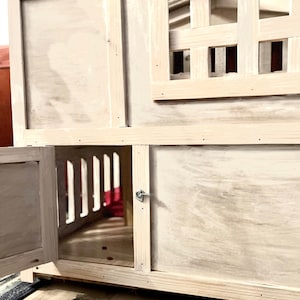 May include: A white wooden cat house with a door that opens and a window with a lattice design. The house is sitting on a tiled floor with two bowls in front of it.