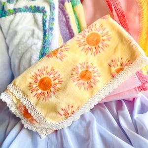 Könnte beinhalten: Ein Stapel weicher Decken in verschiedenen Farben. Die oberste Decke ist gelb mit einem sich wiederholenden orangefarbenen Blumenmuster und weißer Spitzenborte. Weitere Decken sind rosa, weiß und lila, mit Häkel- oder Spitzeneinfassungen.